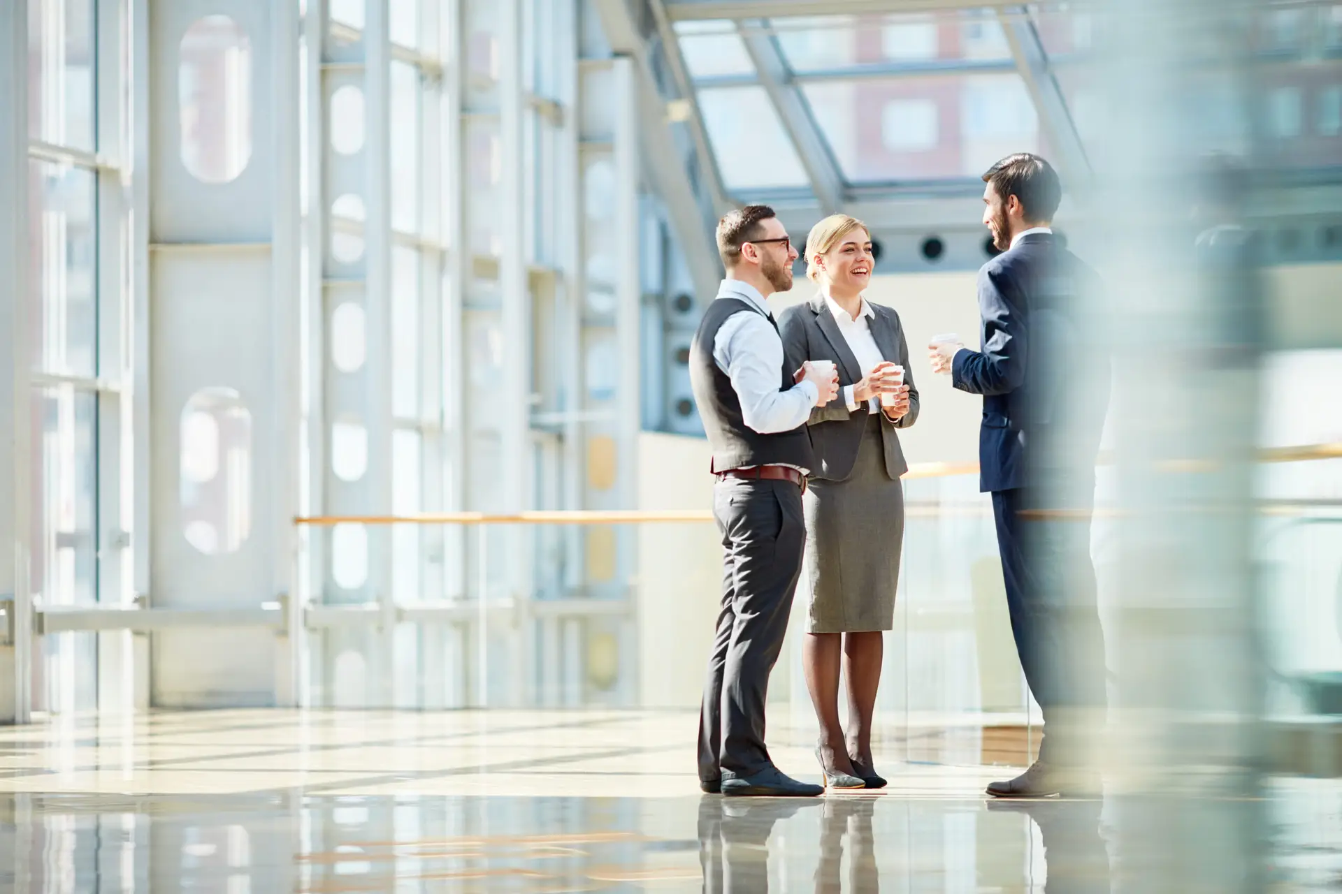 Group of modern business people chatting during coffee break standing in sunlit glass hall of office building
