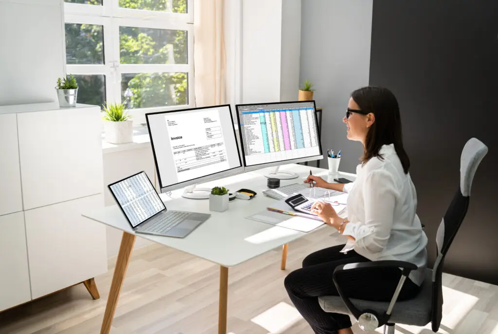 Accountant working at a desk with dual monitors displaying an invoice and spreadsheet, and a laptop