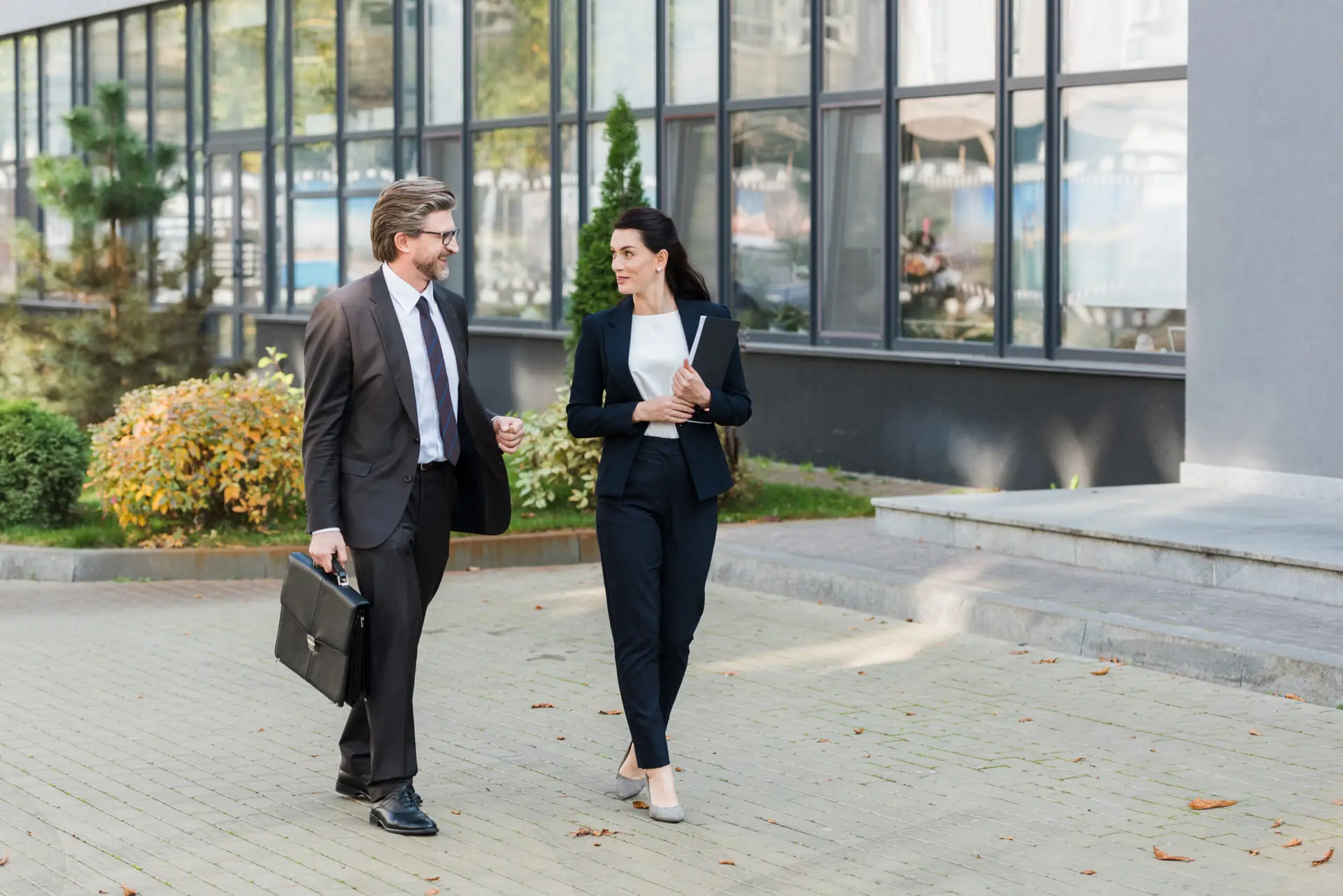 Two business professionals, a man and a woman, walk towards the camera and converse outside a modern office building