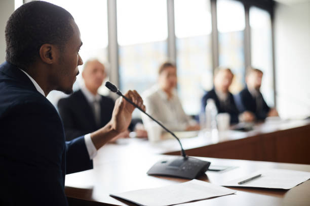 Man in suit speaking into microphone at a conference table with blurred figures in background