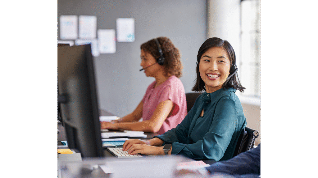 A smiling woman wearing a headset works at a computer in a call center, with another woman in the background